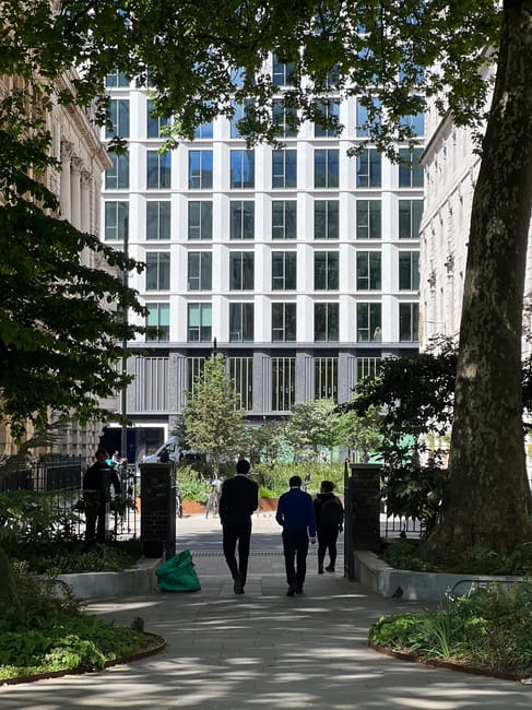 A neatly maintained garden area featuring a paved stone patio with a decorative wrought iron bench positioned centrally in front of a white building with a window and small wrought iron balcony. The g