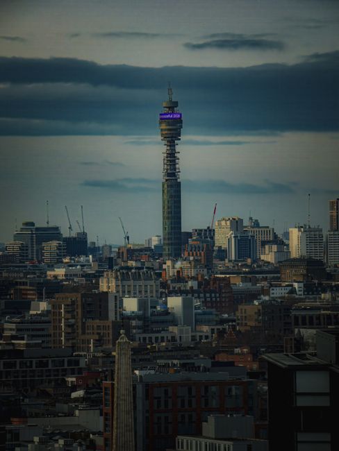 A tall, modern telecommunications tower with a distinctive, multi-tiered cylindrical structure, featuring large glass windows and a prominent blue BT sign near the top. The tower is partially obscured